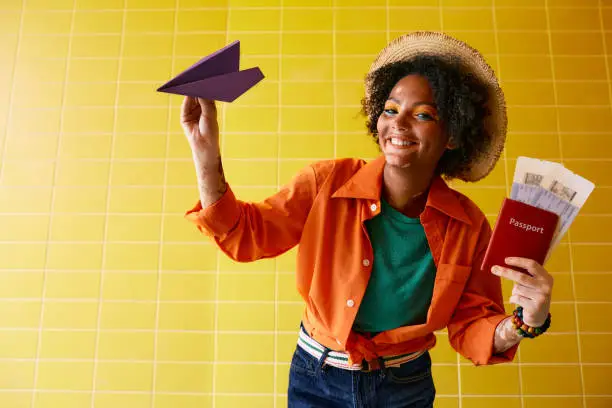 Air travel concept. Smiling multi-ethnic woman with air tickets and international passport throws paper airplane simulating air traveling, over yellow wall background
