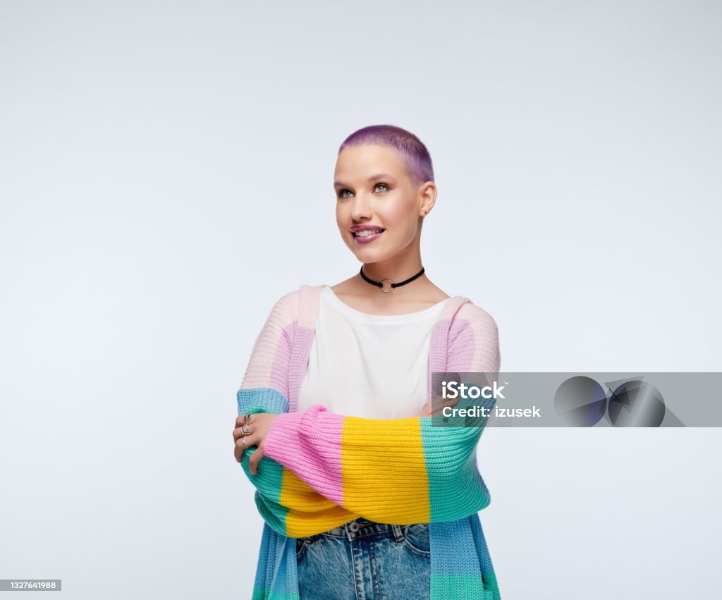 Woman with short purple hair wearing rainbow cardigan Cheerful young woman wearing rainbow cardigan, white shirt and jeans, standing with arms crossed and looking away. Studio portrait on white background. Looking Away Stock Photo