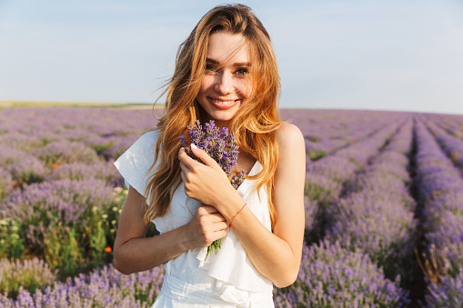 Photo of happy young woman in dress holding bouquet with flowers while walking outdoor through lavender field in summer