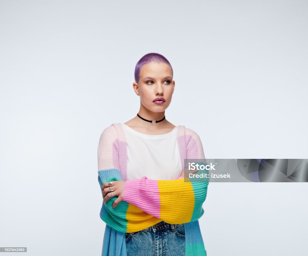 Woman with short purple hair wearing rainbow cardigan Pensive young woman wearing rainbow cardigan, white shirt and jeans, standing with arms crossed and looking away. Studio portrait on white background. 20-29 Years Stock Photo