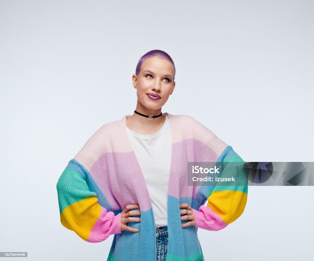 Woman with short purple hair wearing rainbow cardigan Cheerful young woman wearing rainbow cardigan, white shirt and jeans, standing with hands on hips and looking away. Studio portrait on white background. Curiosity Stock Photo