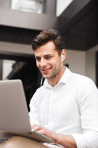 Portrait of a cheerful young formal dressed man in earphones working on laptop computer while sitting outdoors