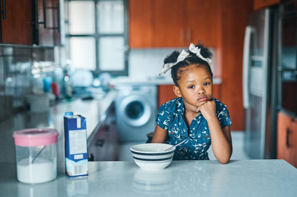 Shot of an adorable little girl sitting alone at the kitchen counter and looking bored before eating breakfast Do you really expect me to eat this every morning? little black girl hairstyle stock pictures, royalty-free photos & images