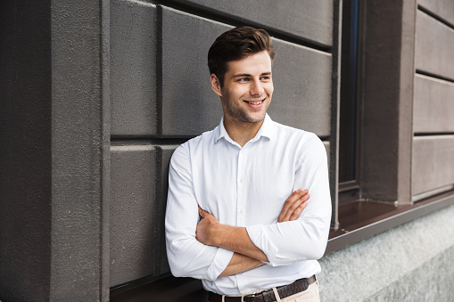 Smiling young male manager formal dressed leaning on a wall outdoors