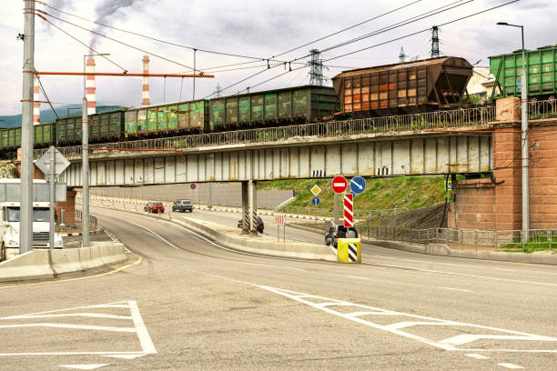 A street in an industrial area of the city. A railway bridge, a train of cars is moving along it. A highway runs under the bridge. Gray-black smoke is coming from the pipes of the enterprise