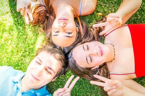 Group of young happy people friends lying in circle on green grass in meadow and relaxing, smiling at camera and showing peace sign, teenagers spending time together outdoors on summer day, top view
