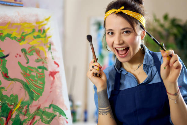 istock Joyful female artist smiling at camera, holding paintbrushes with her hands in paint while creating painting at home studio workshop 1327671228