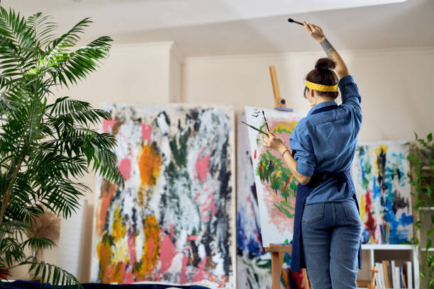 istock Back view of female artist raised her hand with paintbrush in it while creating a new painting on canvas, standing at home studio workshop 1327671237