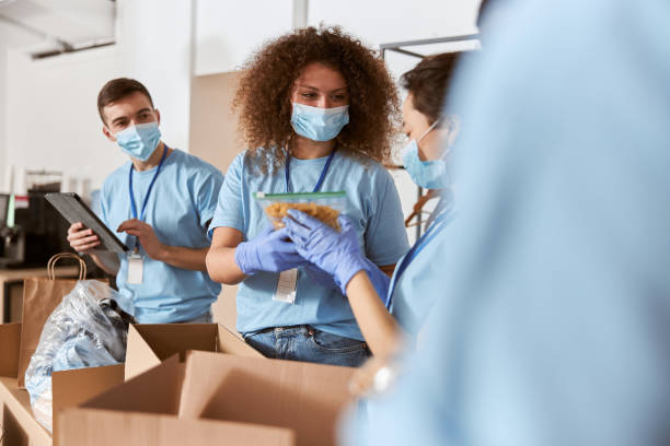 istock Busy volunteers wearing blue uniform, protective masks and gloves sorting donated pack of pasta and other food items while working in charitable foundation 1327672140