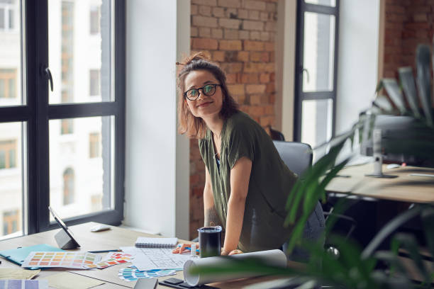 interior designer o architetto in abbigliamento casual con una acconciatura disordinata sorridente alla telecamera mentre lavora con un progetto e campioni di colore per un nuovo progetto, in piedi nel suo ufficio di giorno - architect women blueprint eyewear foto e immagini stock