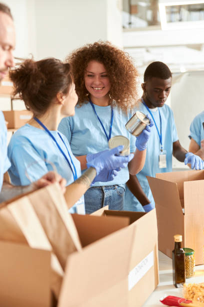 voluntarios alegres en la clasificación de guantes de protección, empacando alimentos enlatados en cajas de cartón, trabajando juntos en el proyecto de donación en interiores - homelessness men white black fotografías e imágenes de stock