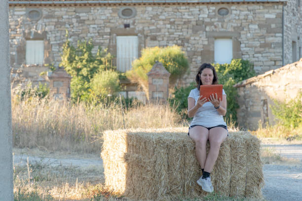 wiejska scena z brunetką latina kobieta konsultuje się z tabletem siedzącym na belce słomy z kamiennym domem w tle - farmer rural scene laptop computer zdjęcia i obrazy z banku zdjęć