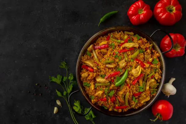 A traditional Chicken paella on dark background with tomatoes and parsley, made of rice with chicken and vegetables in a pan. view from above, flat lay. Copy space
