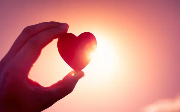 Hand holding heart at sunset against a sun.