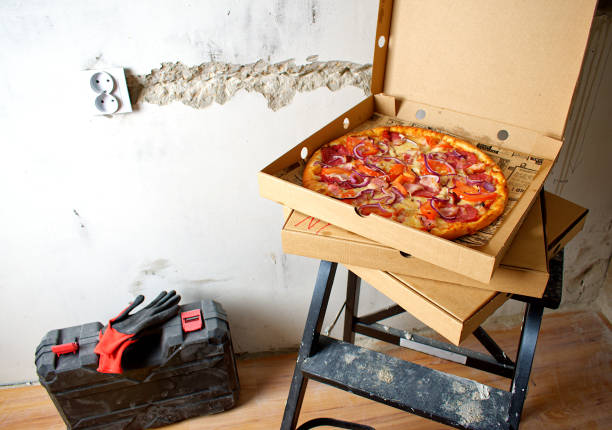 istock Pizza for lunch on the stairs. Take a break from work. Black ladder, hard hat, ear protectors, protective gloves and tool case against the background of a concrete wall. Renovation concept. 1327692620