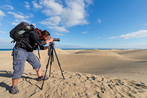 Taking pictures in the desert.\nlong haired photographer taking photo with tele lens on tripod in desert by beach.\nCanary Islands, Gran Canaria