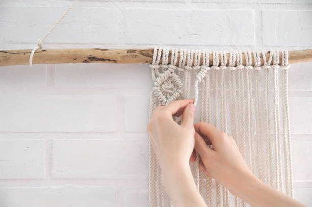 woman knits weaving macrame pinned on a wooden stick near a white brick wall woman knits weaving macrame pinned on a wooden stick near a white brick wall macrame stock pictures, royalty-free photos & images