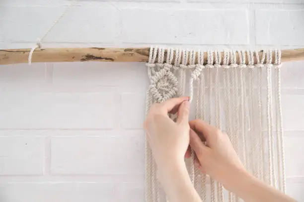 Photo of woman knits weaving macrame pinned on a wooden stick near a white brick wall