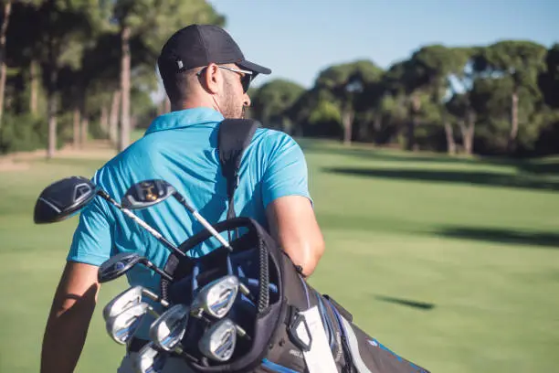 Young golfer walking by carrying golf bag in golf course.
