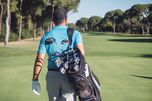 Young golfer walking by carrying golf bag in golf course.