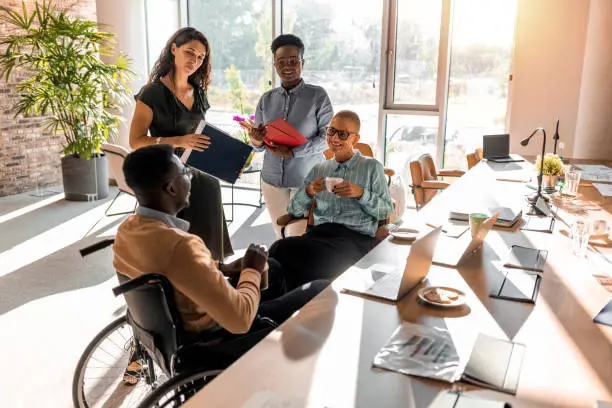 Photo of diverse team looks happy in conversation. They are sitting and standing around with laptops in front of them.