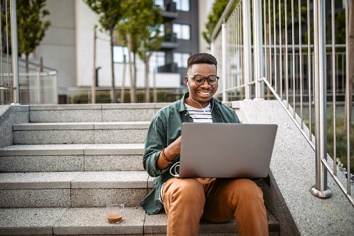 Young man sitting on the stairs in a front of the office building, working using laptop