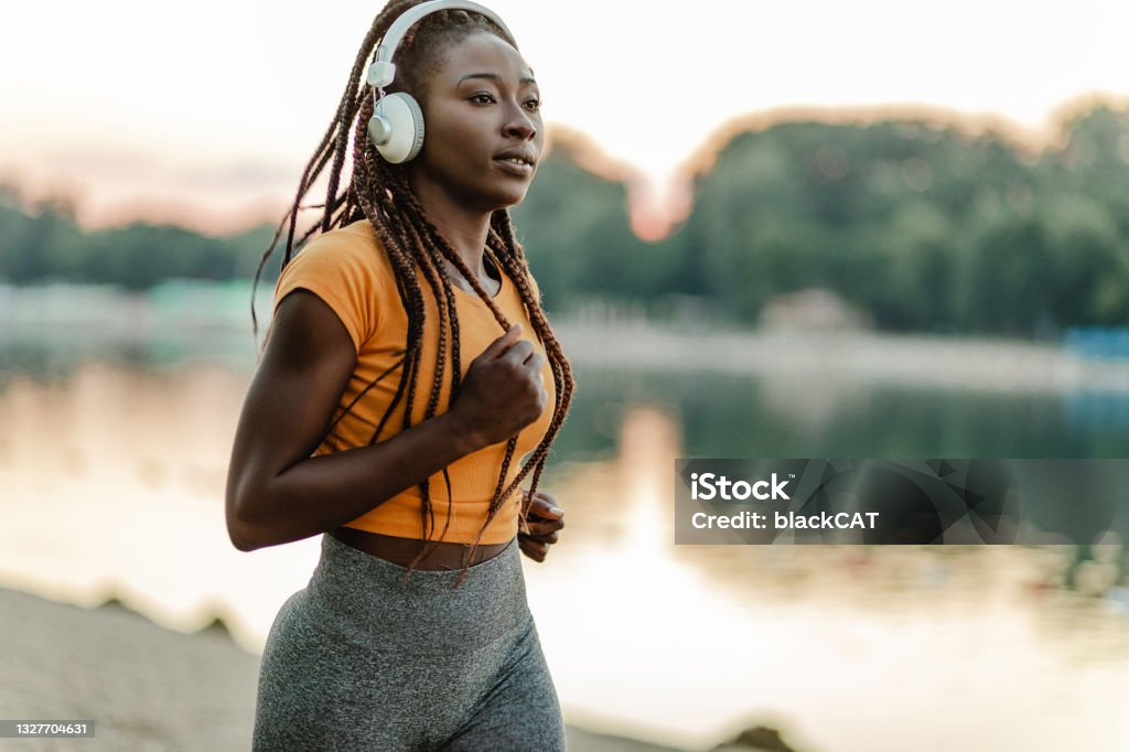 Young African American woman is jogging by the river African American woman doing sport by the river and listening music with headphones Running Stock Photo