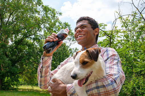 Young smiling cheerful African American man holding Jack Russell terrier dog and drinking cola from bottle outdoors. Walking in park in summer, low angle view