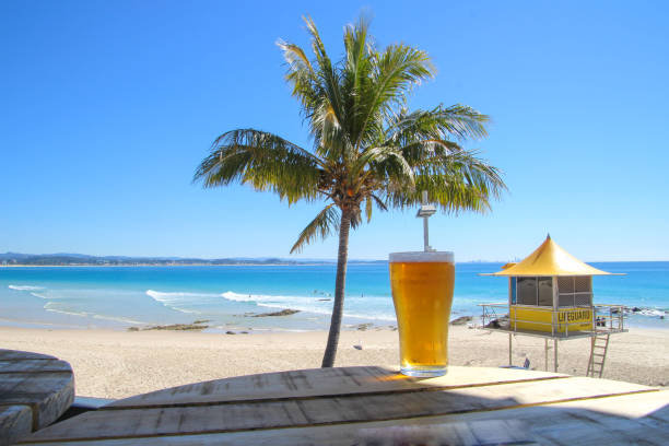 ヤシの木とライフガードタワーと青い海を見下ろす木材テーブルの上のビールの冷�たいガラス。 - gold coast australia lifeguard sea ストックフォトと画像