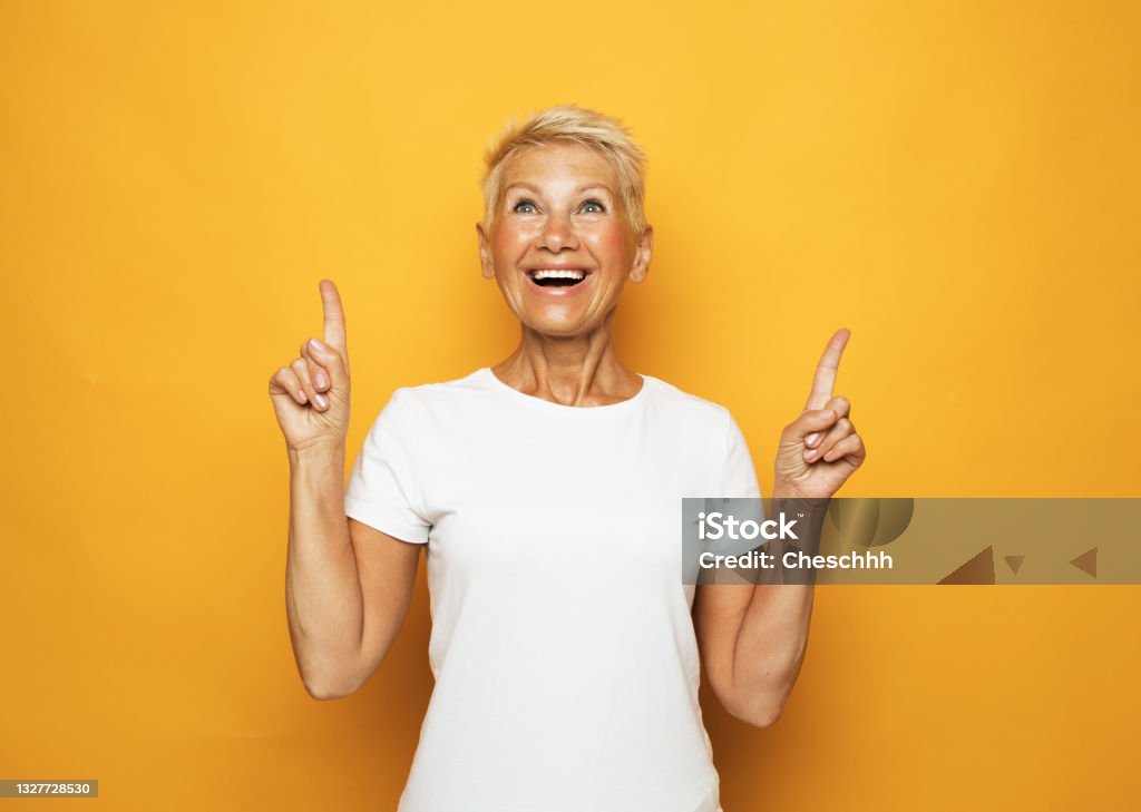 Senior woman with short hair pointing with fingers and raised arms lifestyle, emotion and old people concept: Senior woman with short hair wearing white t-shirt amazed and surprised looking up and pointing with fingers and raised arms. White People Stock Photo