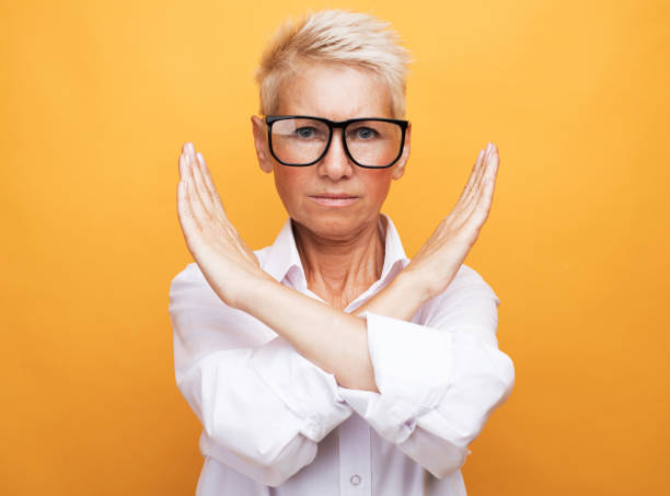 Senior woman rejection expression crossing arms doing negative sign lifestyle, emotion and old people concept: Senior woman with short gray hair wearing white shirt and glasses rejection expression crossing arms doing negative sign over yellow background refusing stock pictures, royalty-free photos & images