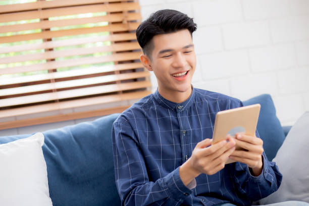 istock Young attractive asian man resting using browsing tablet computer on sofa at home, happy male sitting on couch relax reading digital gadget at house, communication and lifestyle concept. 1327730922