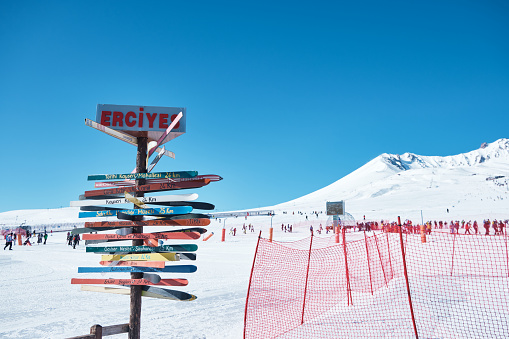 Direction signpost at Erciyes ski resort with distancies to different cities against snow covered peak at background. Kayseri, Turkey - February 2021