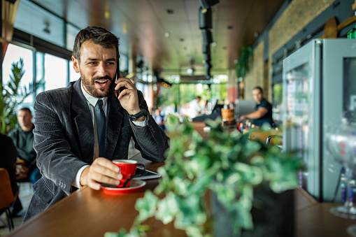 Young Confident Businessman is Sitting in a Coffee Shop, Drinking a Hot Cup of Coffee and Talking with his Business Partners and Working on Digital Tablet. Handsome Man in Business Wear is Having a Coffee Break in Local Restaurant and has a Conversation Using a Phone.