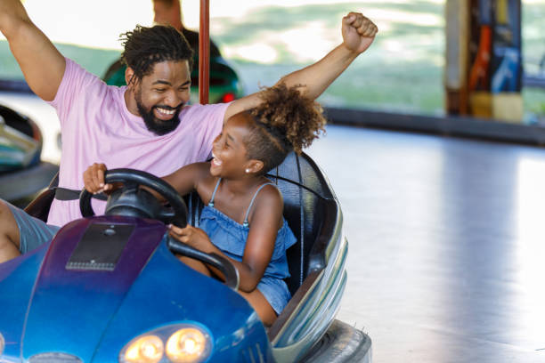 An African-American Family is Enjoying in Driving Bumper Car in Amusement Park. Father and Daughter of African-American Ethnicity are Spending a Wonderful Day While Driving in Bumper Car in Luna Park. amusement park stock pictures, royalty-free photos & images