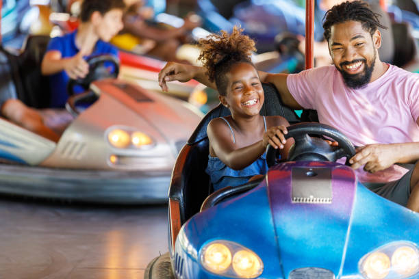 African-American Father is Driving in Bumper Car with his Cute Little Daughter. A Beautiful Little Daughter is Happy to Spend a Wonderful Day with her Happy Father. They are Driving an Electric Car in the Amusement Park. amusement park ride photos stock pictures, royalty-free photos & images