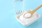 Dish of salt with wooden spoon and glass of water on light blue background