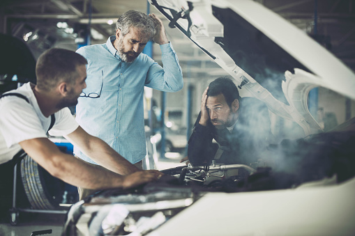 Mid adult customer and mechanics feeling frustrated while the car is smoking in a repair shop.