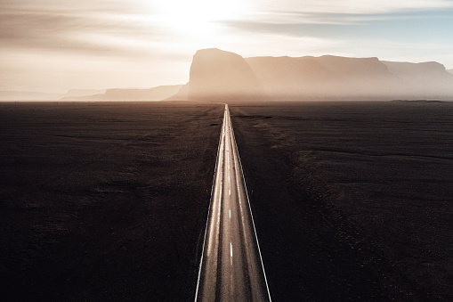 Endless straight road into the sun between black volcanic gravel towards sunset over icelandic mountain range. Aerial drone point of view along the country highway. Toned Image Colors. Southern Iceland, Golden Circle Route, Iceland, Northern Europe.