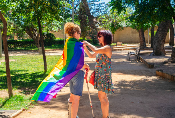An adult woman ties a rainbow flag around the neck of her disabled girlfriend on crutches in a public park. Concept of diversity stock photo