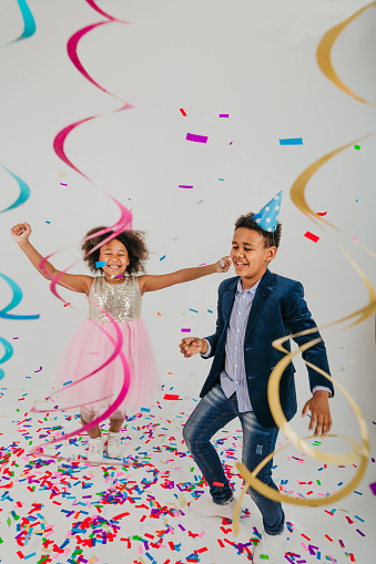 Happy girl and boy in party hats dancing on white background decorated with serpentine and confetti