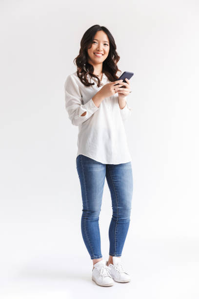 photo pleine longueur d’une femme chinoise brune aux longs cheveux foncés tenant et utilisant un smartphone noir, isolée sur fond blanc en studio - standing photos et images de collection