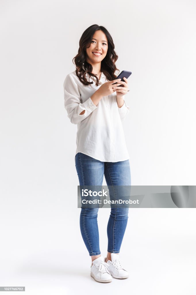 Full length photo of brunette chinese woman with long dark hair holding and using black smartphone, isolated over white background in studio Full length photo of brunette chinese woman with long dark hair holding and using black smartphone isolated over white background in studio Women Stock Photo