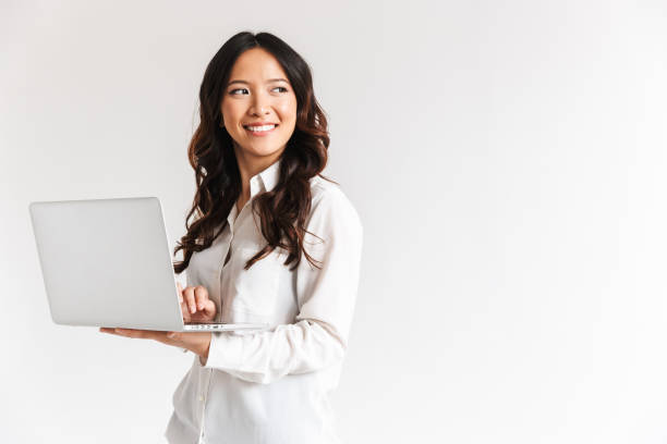 Smiling young asian businesswoman standing Smiling young asian businesswoman standing with laptop computer over white background, looking away asian culture stock pictures, royalty-free photos & images