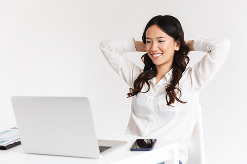 Photo of joyous chinese businesswoman with long dark hair sitting in office and working with documents and laptop isolated over white background