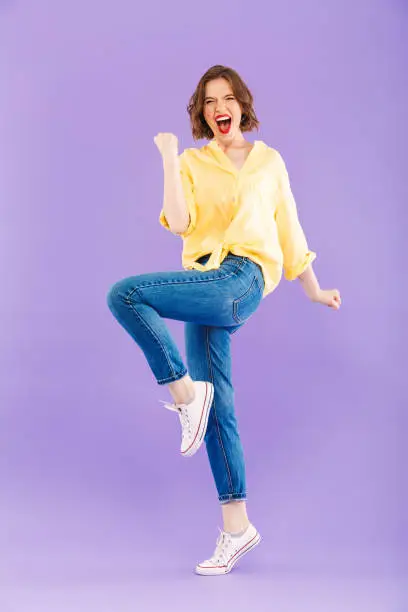 Full length portrait of a cheerful young woman dressed in summer clothes celebrating success over violet background