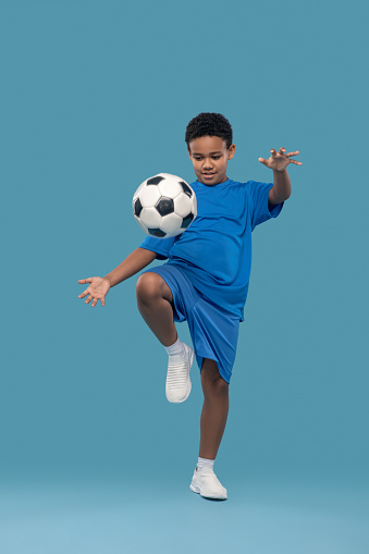 Favorite sport. Dark-skinned persistent trying boy in shorts and tshirt throwing soccer ball with knee up on blue background