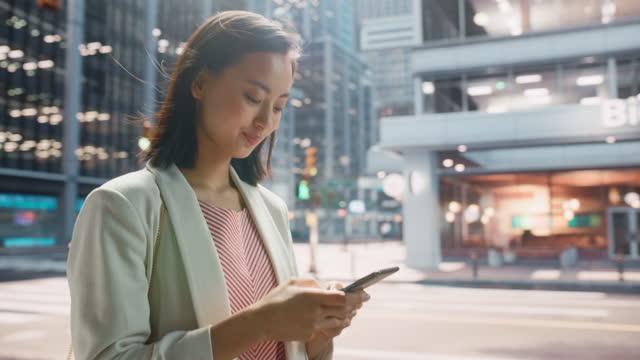 Portrait of an Attractive Japanese Female Walking in Smart Casual Clothes and Using Smartphone on the Urban Street. Manager in Big City Connecting with People Online, Messaging and Browsing Internet.