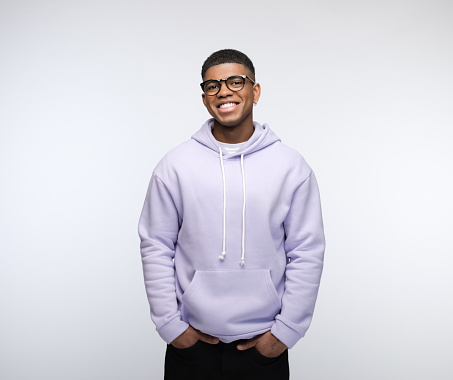 Happy african young man wearing lilac hoodie, standing with hands in pockets and laughing at camera. Studio portrait on white background.