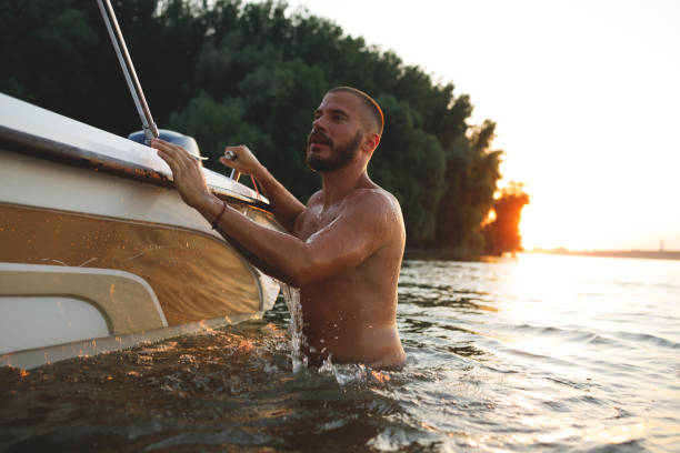 A handsome young man is swimming in the Danube river next to his speed boat, alone at sunset. He enjoys the peace and beauty of nature around the river away from the formal beach.
